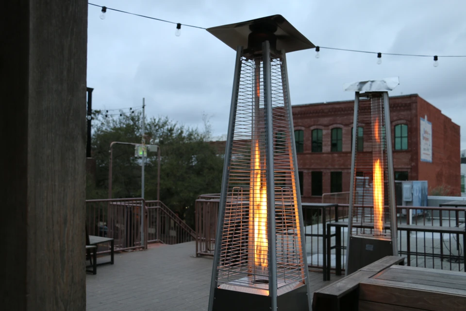 Terrasse extérieure d'établissement avec chauffe-terrasse pyramidal à gaz allumé, diffusant une flamme orange vif à travers structure métallique grillagée